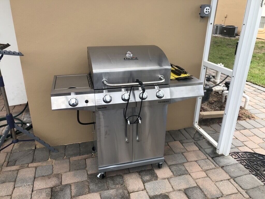 Ein tragbarer Grill aus Edelstahl mit vier Brennern und einem Seitenbrenner, auf einer gepflasterten Terrasse platziert.