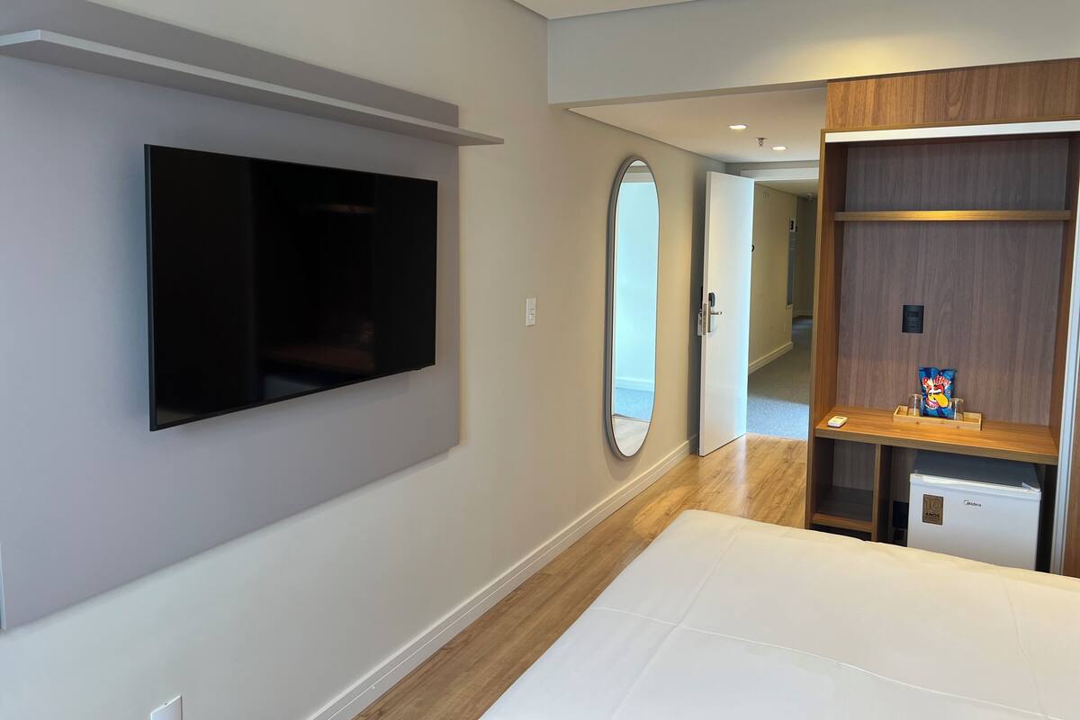 Quarto de hotel moderno com televisão de tela plana na parede, um espelho grande, guarda-roupa de madeira e minibar.