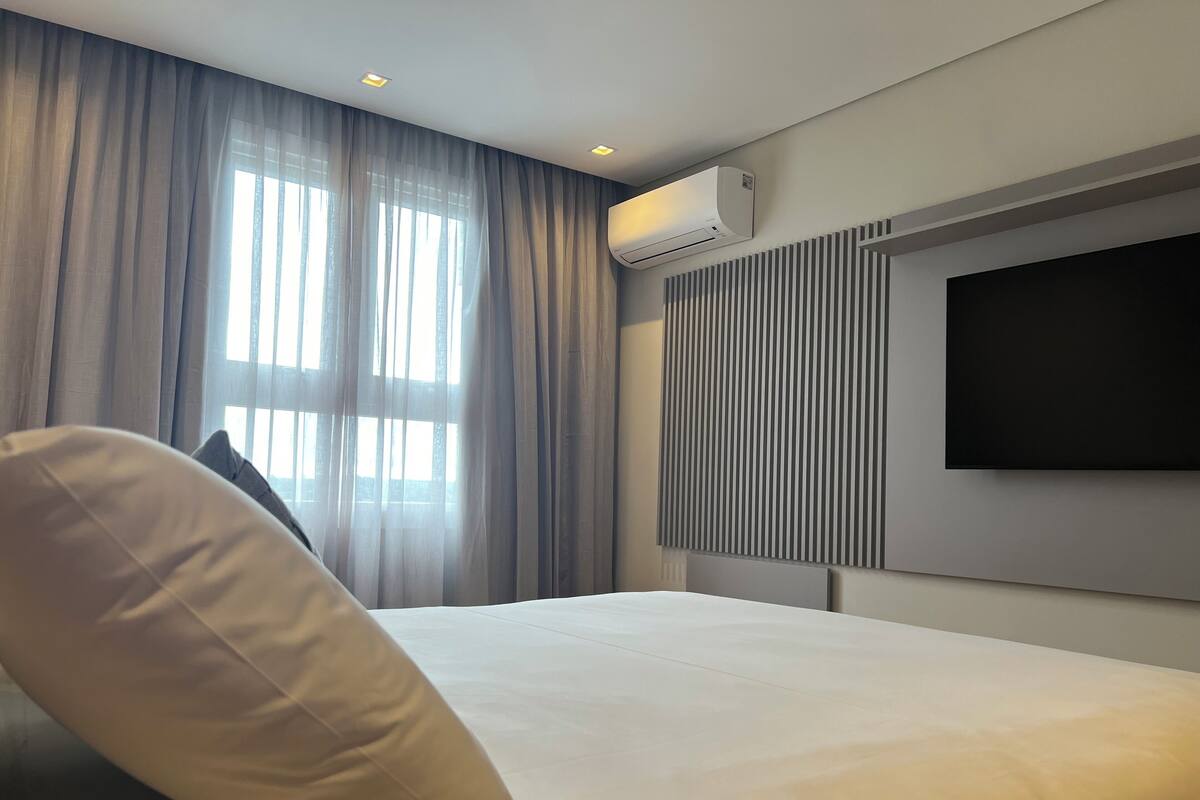 Quarto de hotel moderno com uma cama grande, uma televisão de tela plana na parede e uma janela com cortinas translúcidas.