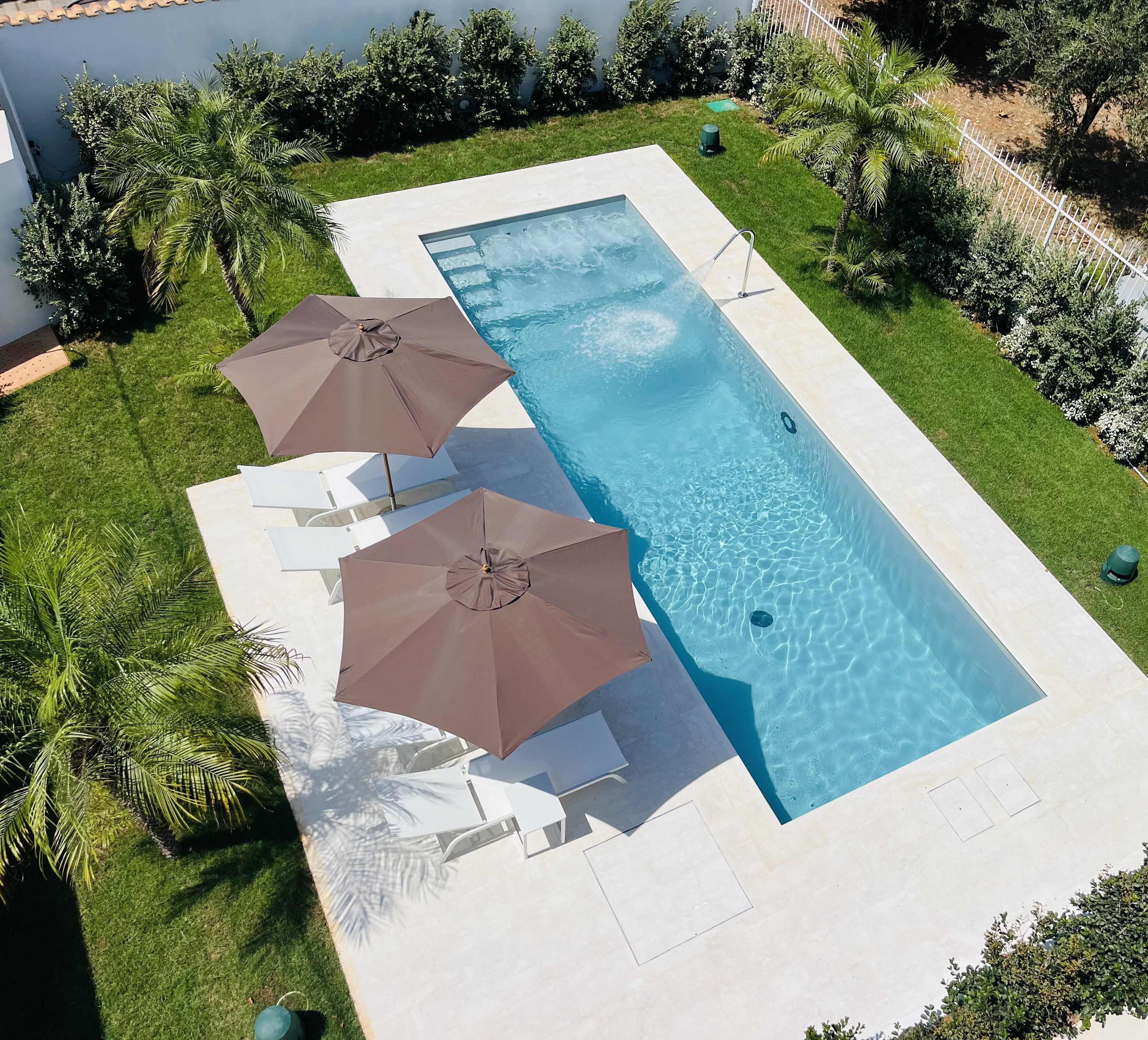 Ein rechteckiger Swimmingpool mit klarem, blauem Wasser, umgeben von einer gepflegten Rasenfläche und Palmen. Zwei Sonnenschirme spenden Schatten über Liegestühlen.