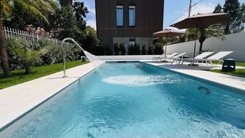 A rectangular swimming pool with a water fountain, surrounded by a white fence, palm trees, and a modern building with large windows.