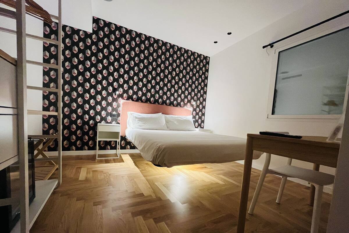 A bedroom with a bed, a desk, a chair, a TV, and a patterned wall.