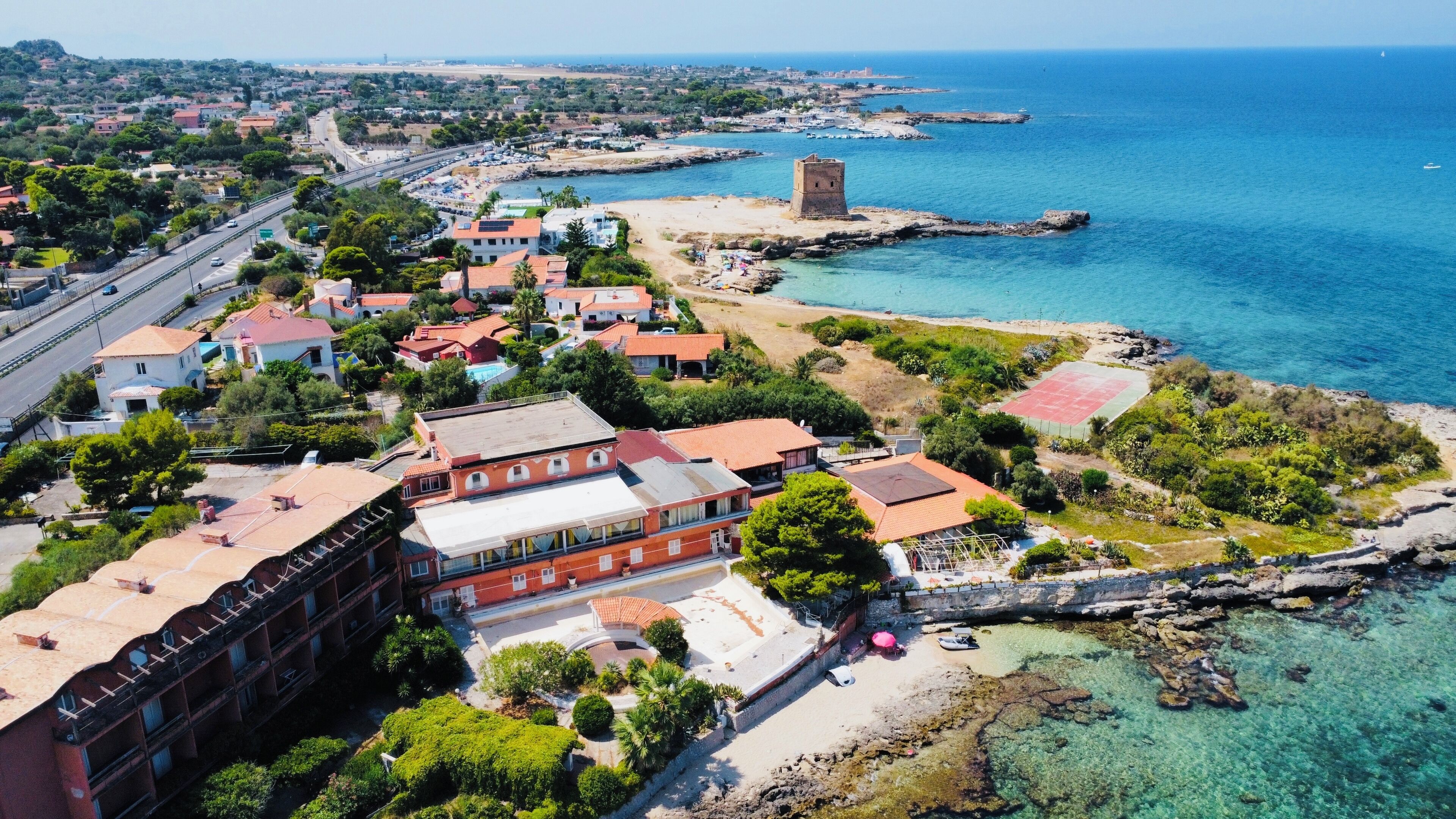 Vista aérea de un pueblo costero con un edificio destacado, mar azul cristalino y costa rocosa.