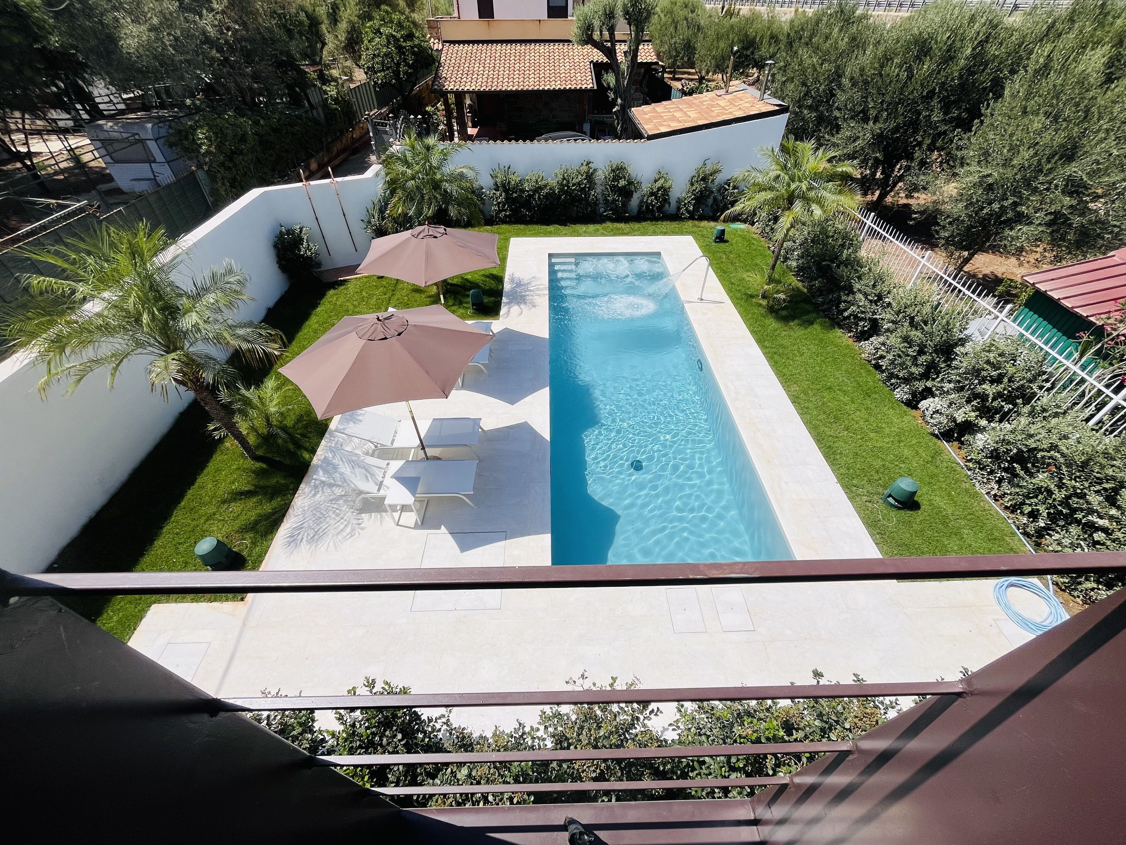 A rectangular swimming pool with a clear blue water surface, surrounded by a white wall and a small grassy area. There are two white lounge chairs and a brown umbrella providing shade.