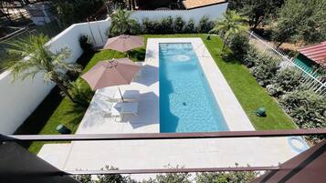 A rectangular swimming pool with a clear blue water surface, surrounded by a white wall and a small grassy area. There are two white lounge chairs and a brown umbrella providing shade.