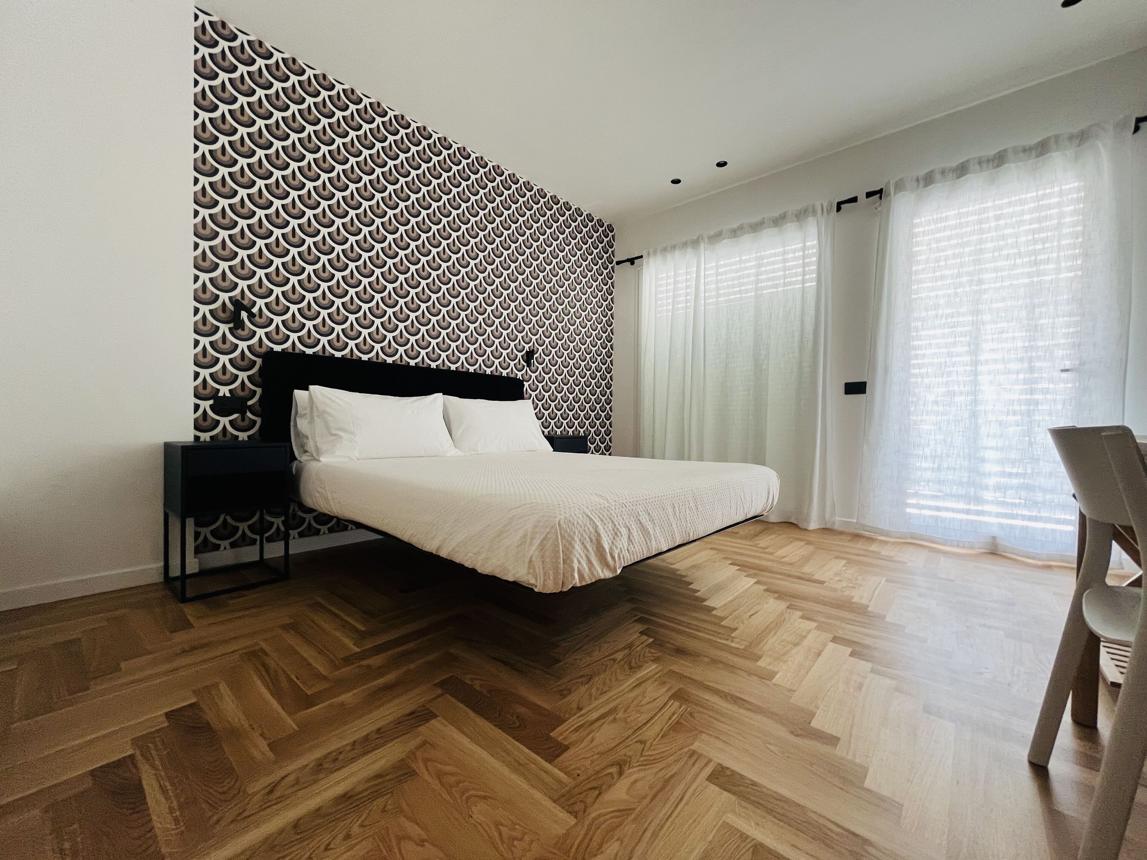 Een moderne slaapkamer met een behangwand, een houten visgraatvloer, een bed met witte beddengoed en een stoel.