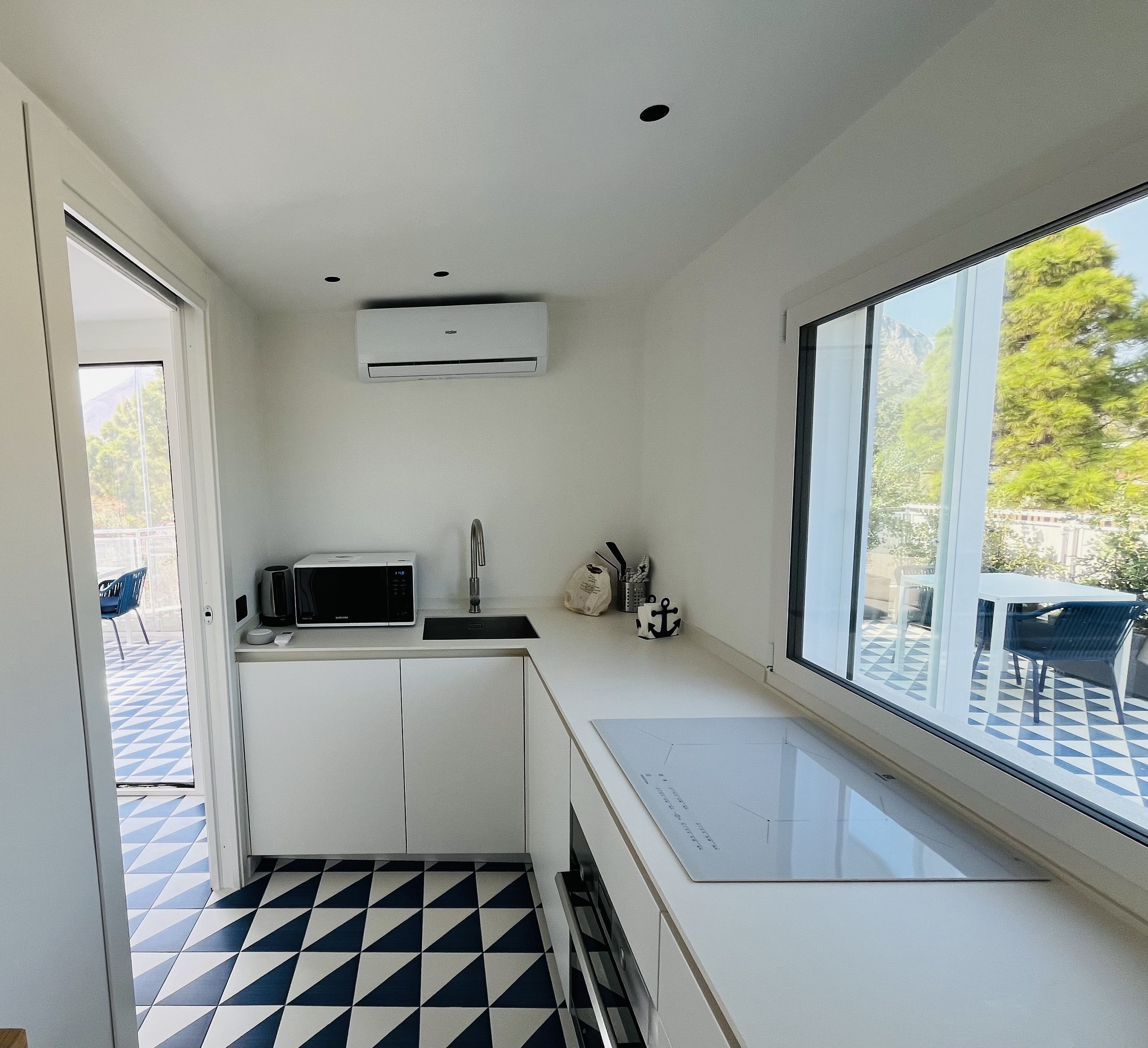 Una cocina moderna con armarios blancos, un microondas y un fregadero. Hay una ventana con vista a árboles y un patio exterior.