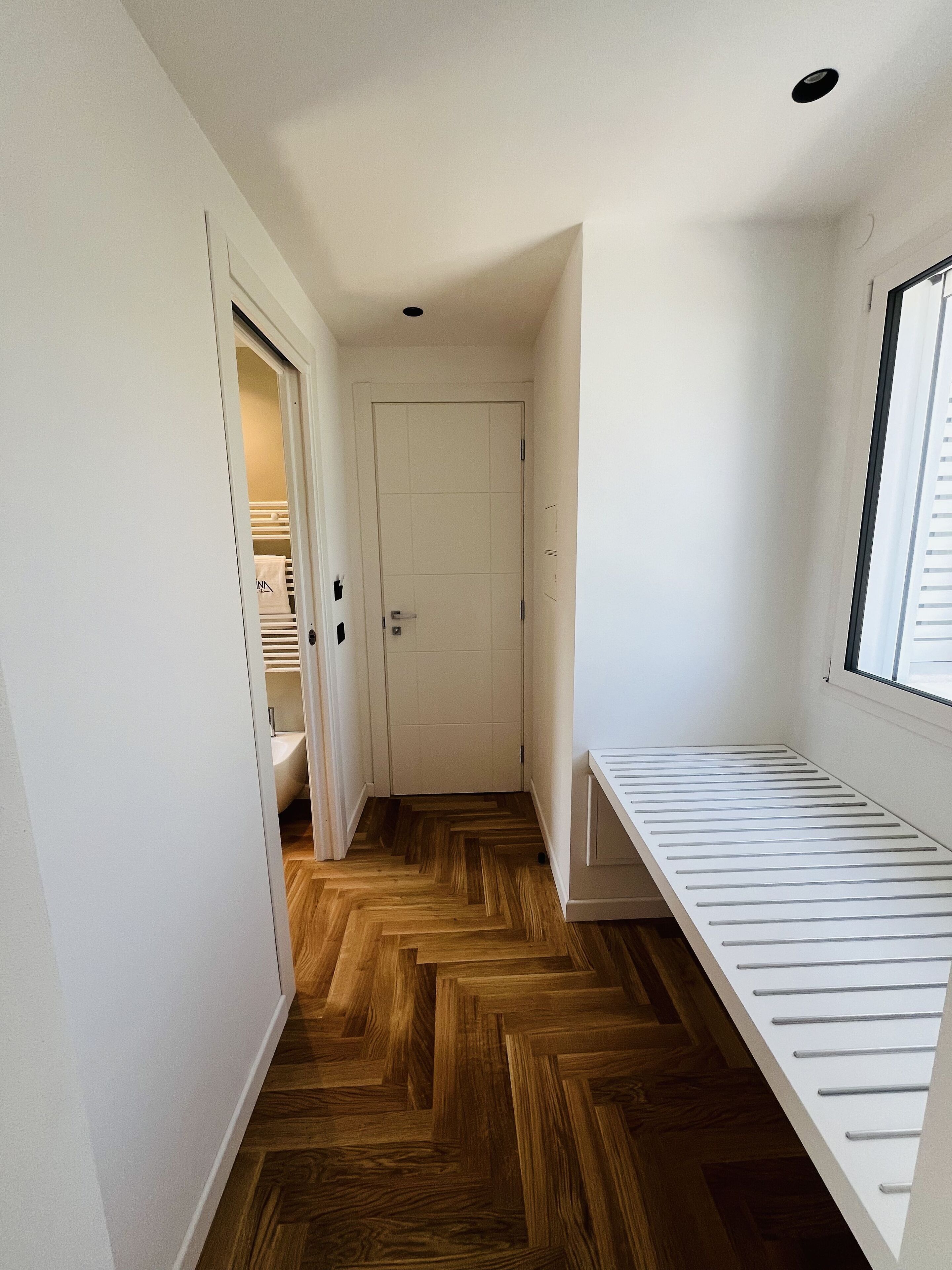 Ein schmaler Flur mit Holzdielenmuster, einer weißen Tür und einem Fenster auf der rechten Seite.