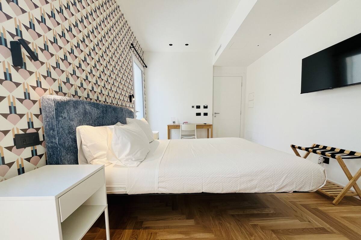 A modern bedroom with a geometric-patterned wall, a bed with white bedding, a wooden floor, a desk, a chair, a TV mounted on the wall, and a small ironing board.