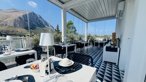 Μια βεράντα με τραπέζια και καρέκλες, ένα λευκό ταβάνι και θέα στο βουνό.