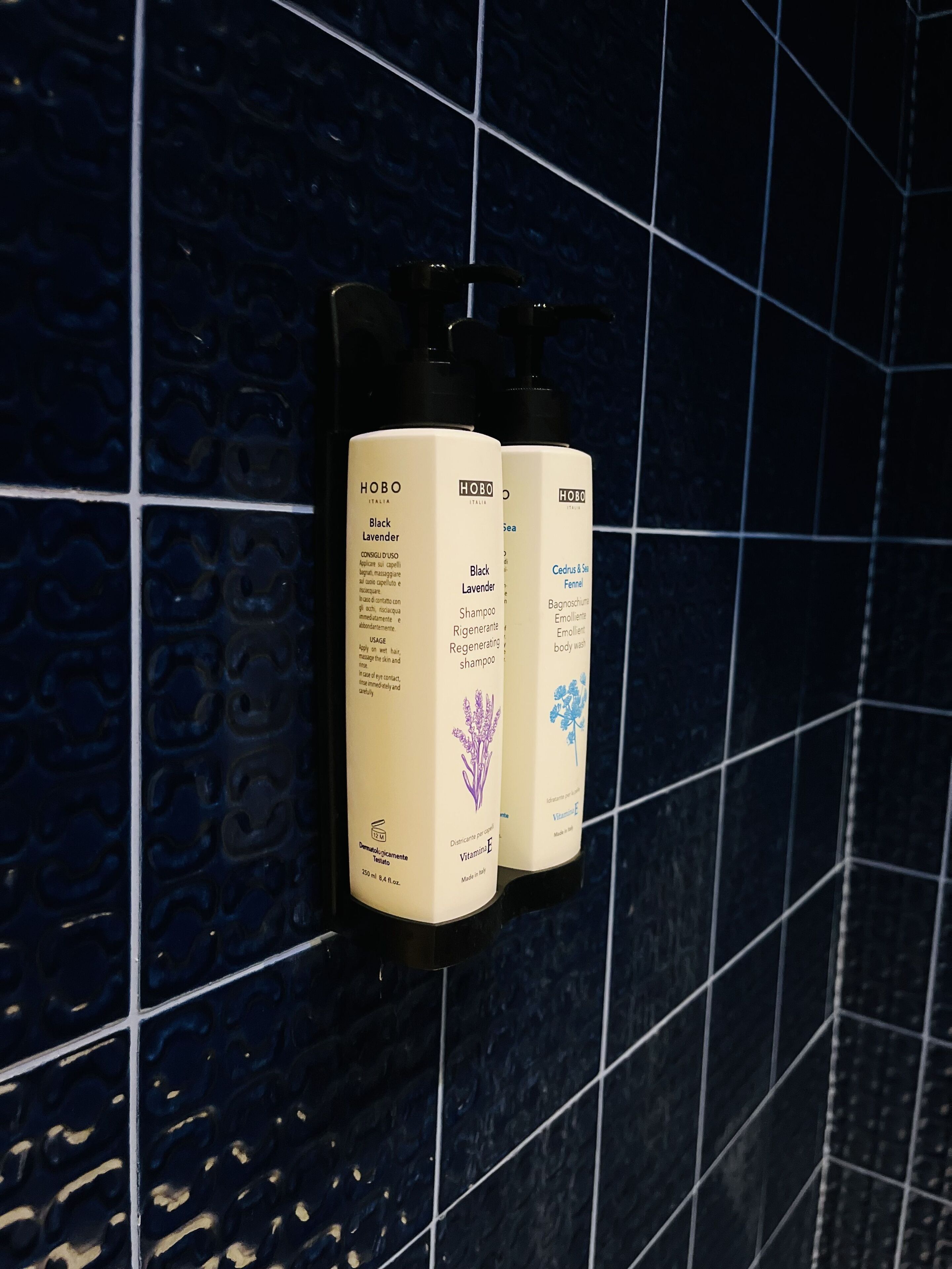 Drie flessen HOBO Italy Black Lavender shampoo bevestigd aan een donkerblauwe betegelde muur.