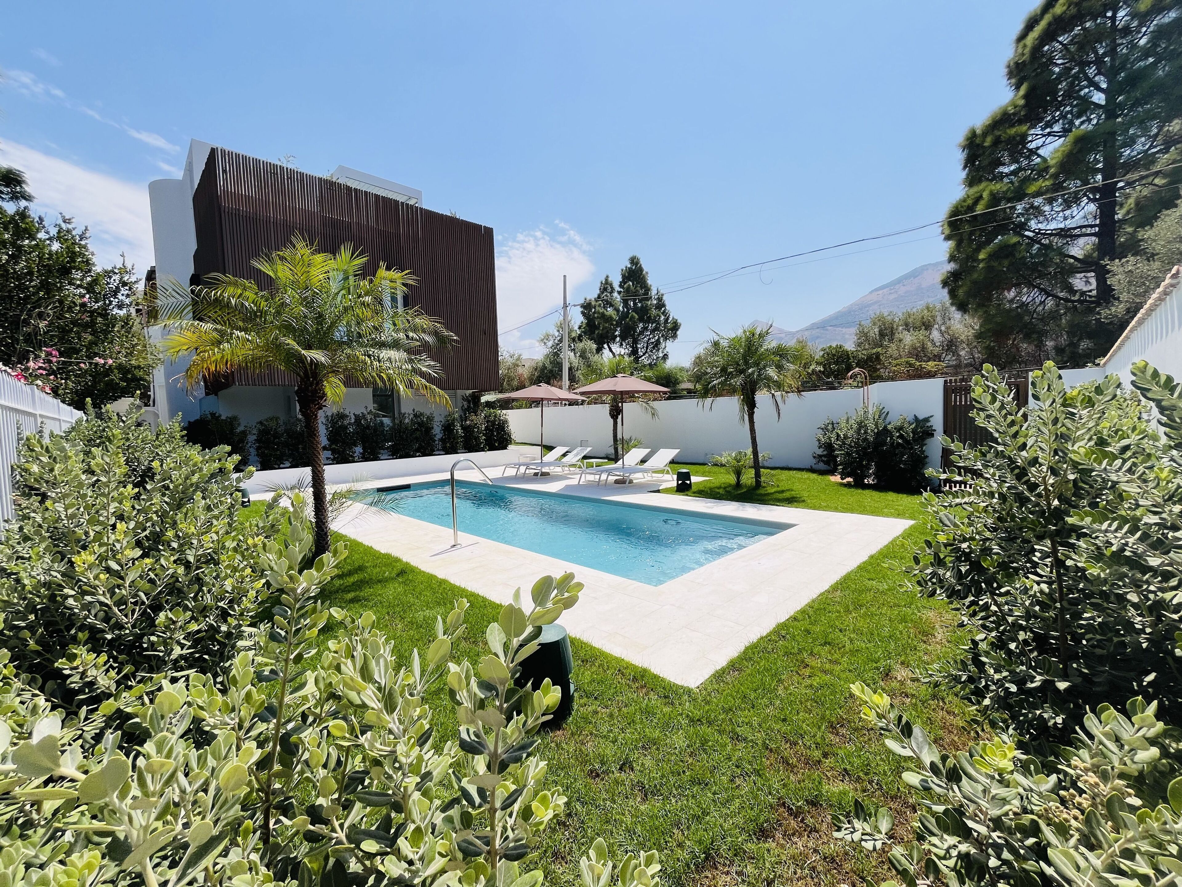 Un área de piscina moderna al aire libre con una piscina rectangular, sillas de descanso y una pérgola.