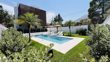 Un área de piscina moderna al aire libre con una piscina rectangular, sillas de descanso y una pérgola.