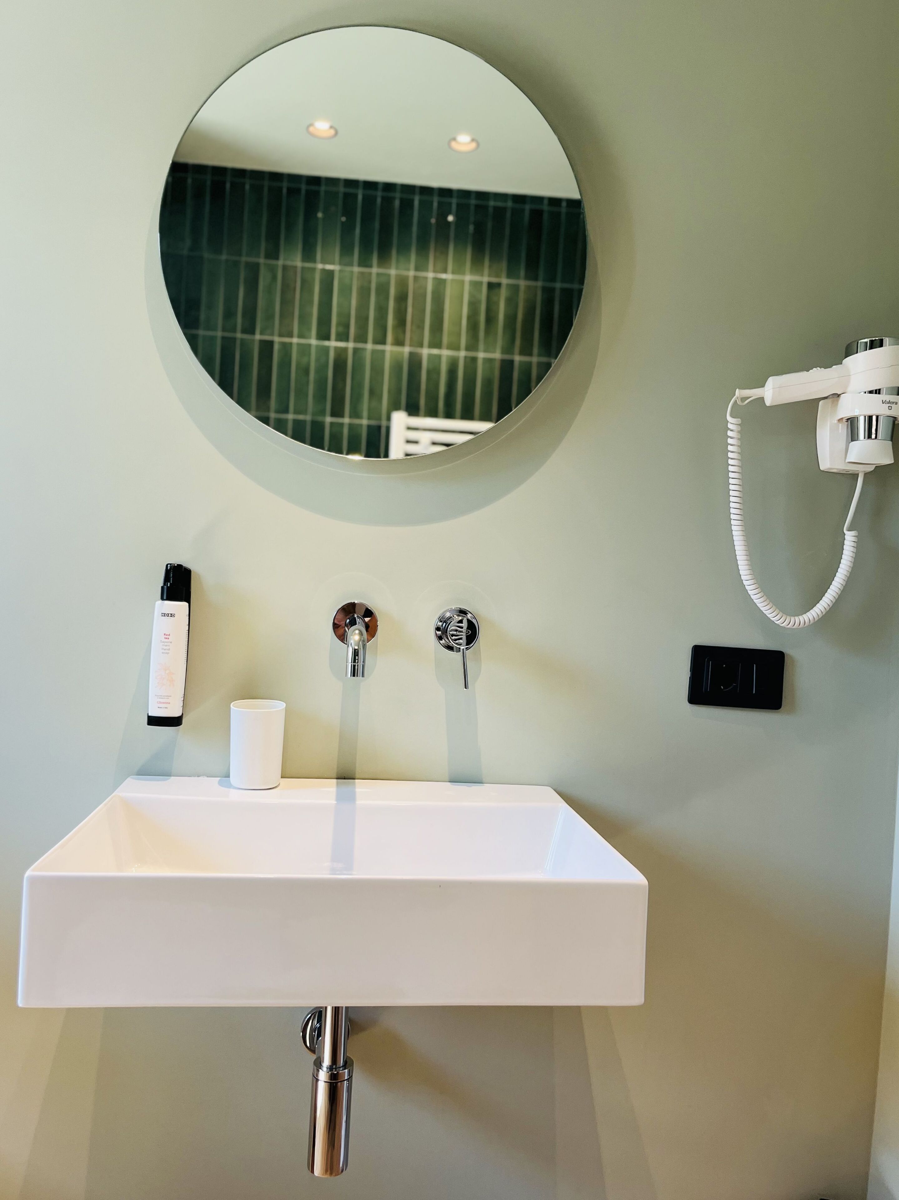 Un bagno con un lavandino bianco, uno specchio rotondo, un soffione a mano e una bottiglia di shampoo.
