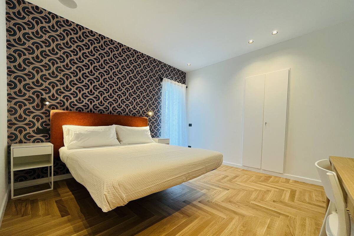 A bedroom with a patterned wallpaper, a wooden floor, a bed with white bedding, a nightstand, and a wardrobe.