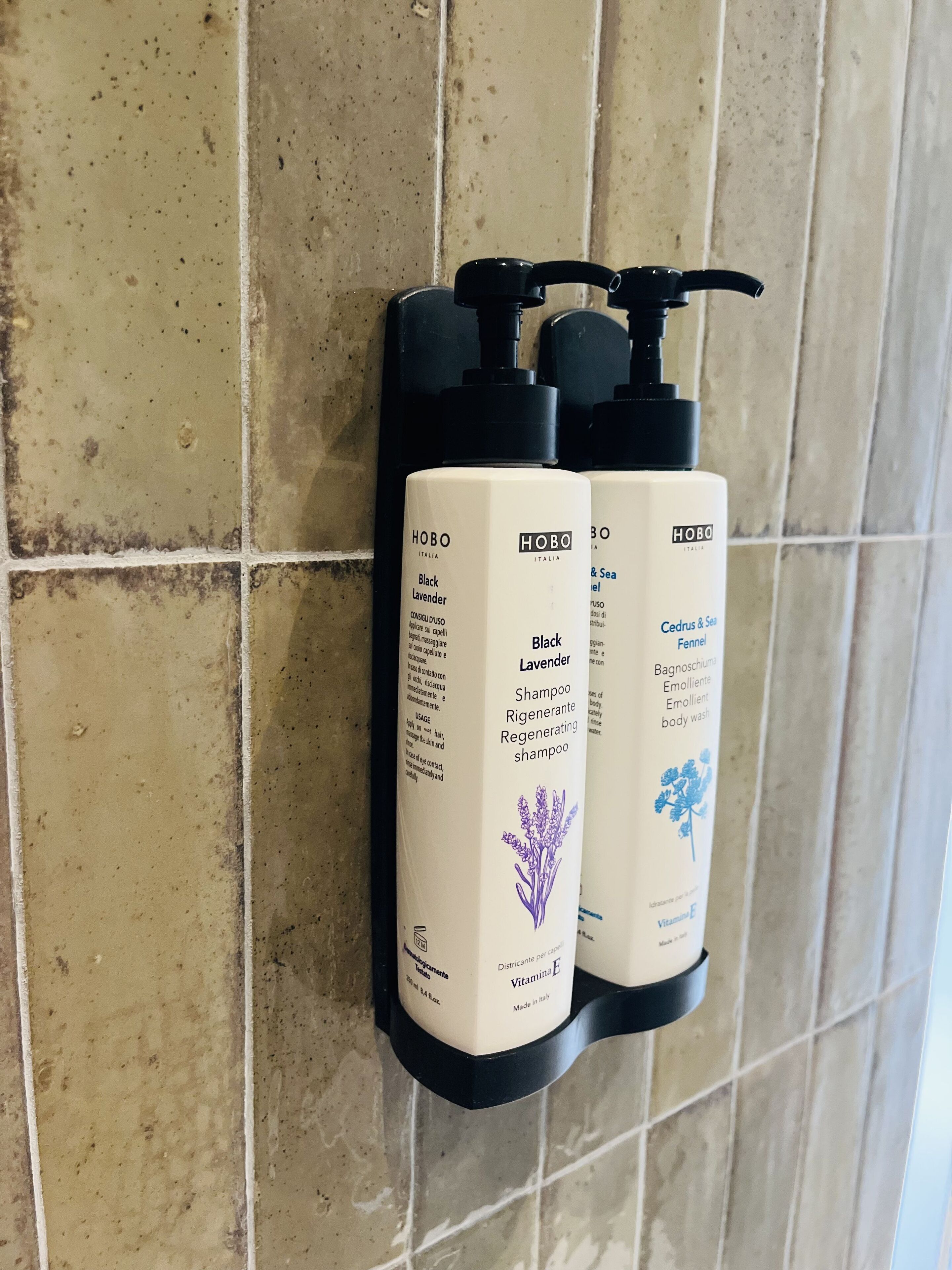 Two HOBO Italy Black Lavender Shampoo dispensers mounted on a tiled wall.