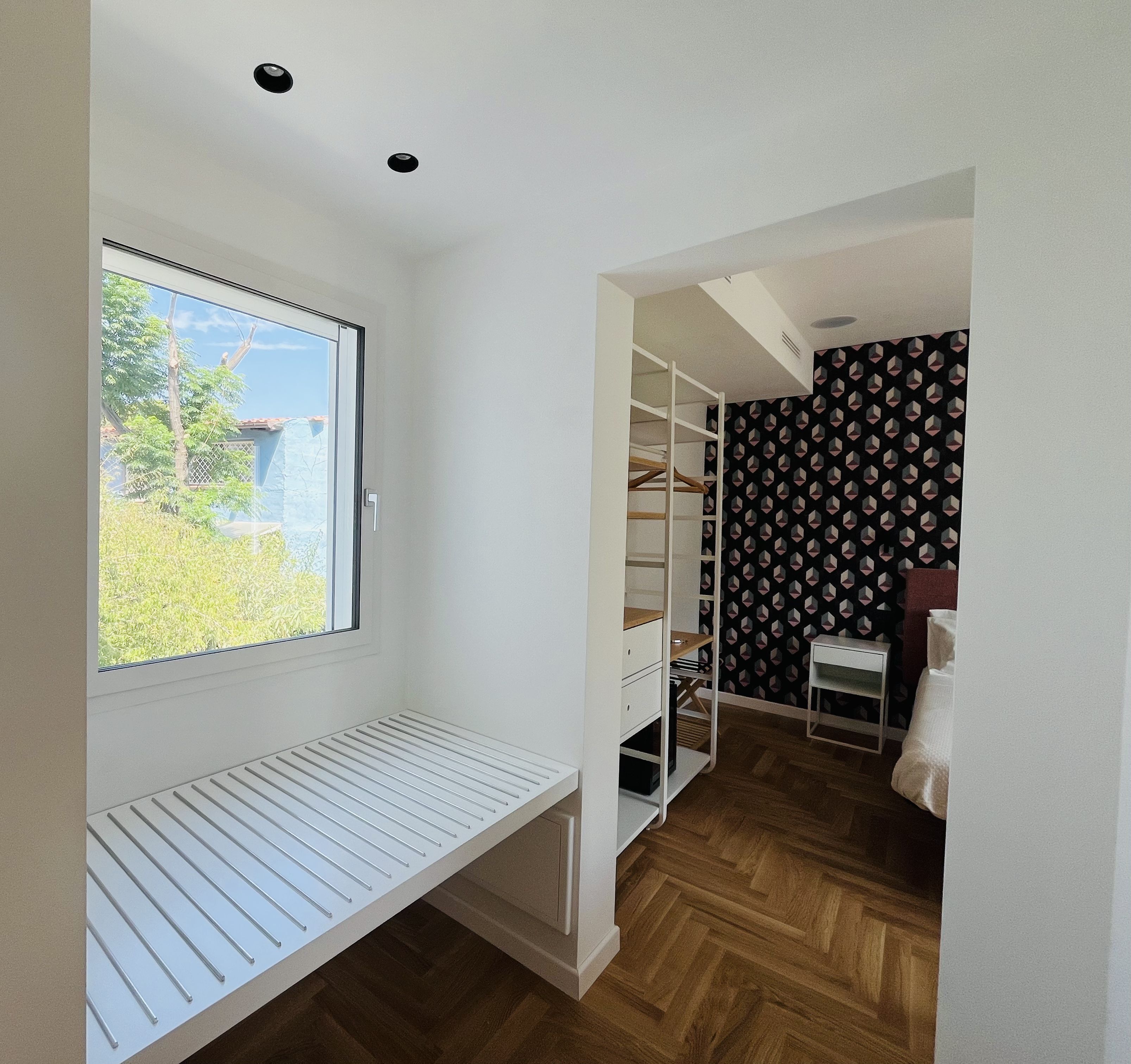 A small, well-lit room with a window, a built-in bench, a wooden floor, and a closet with a patterned wallpaper.