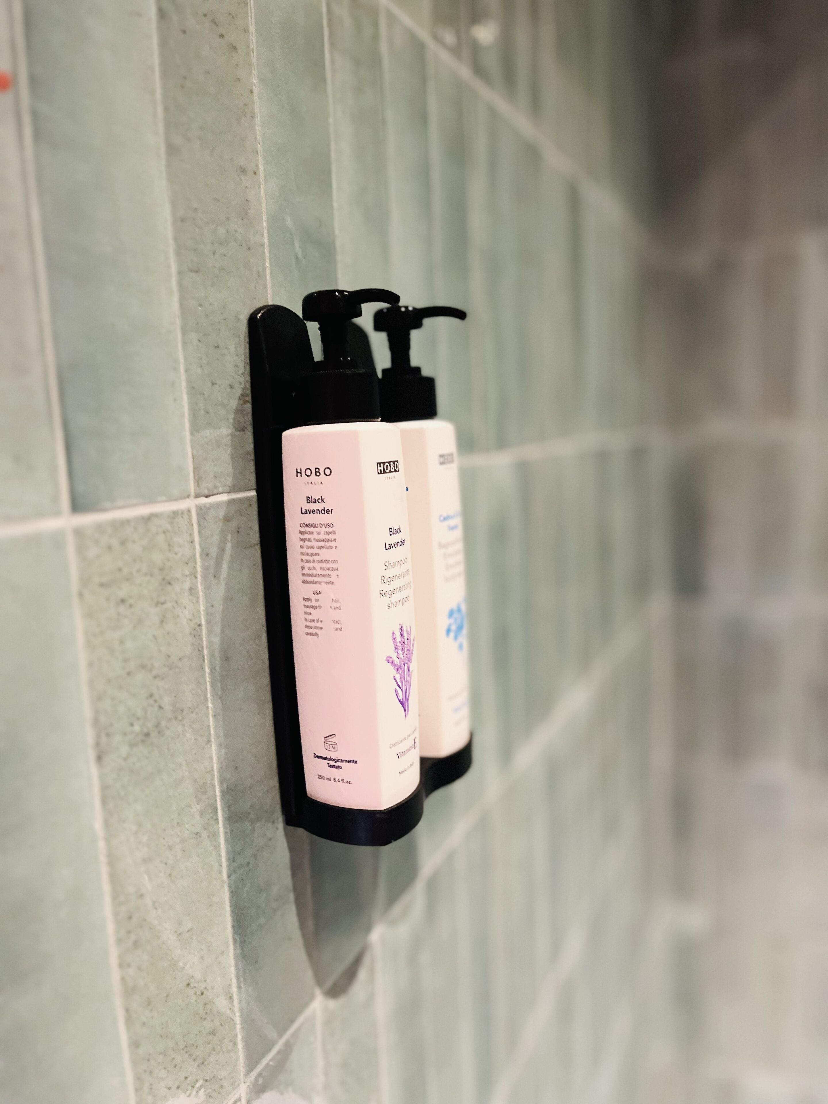 Zwei schwarze und pinke Flaschen HOBO Black Lavender Shampoo und Conditioner auf einer gefliesten Wand.