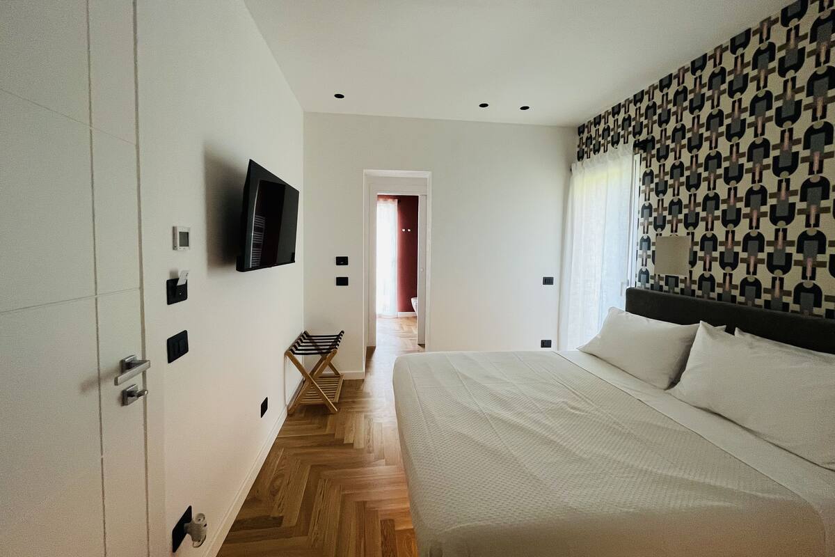 A modern hotel room with a large bed, a flat-screen TV mounted on the wall, a wooden floor, and a patterned headboard.