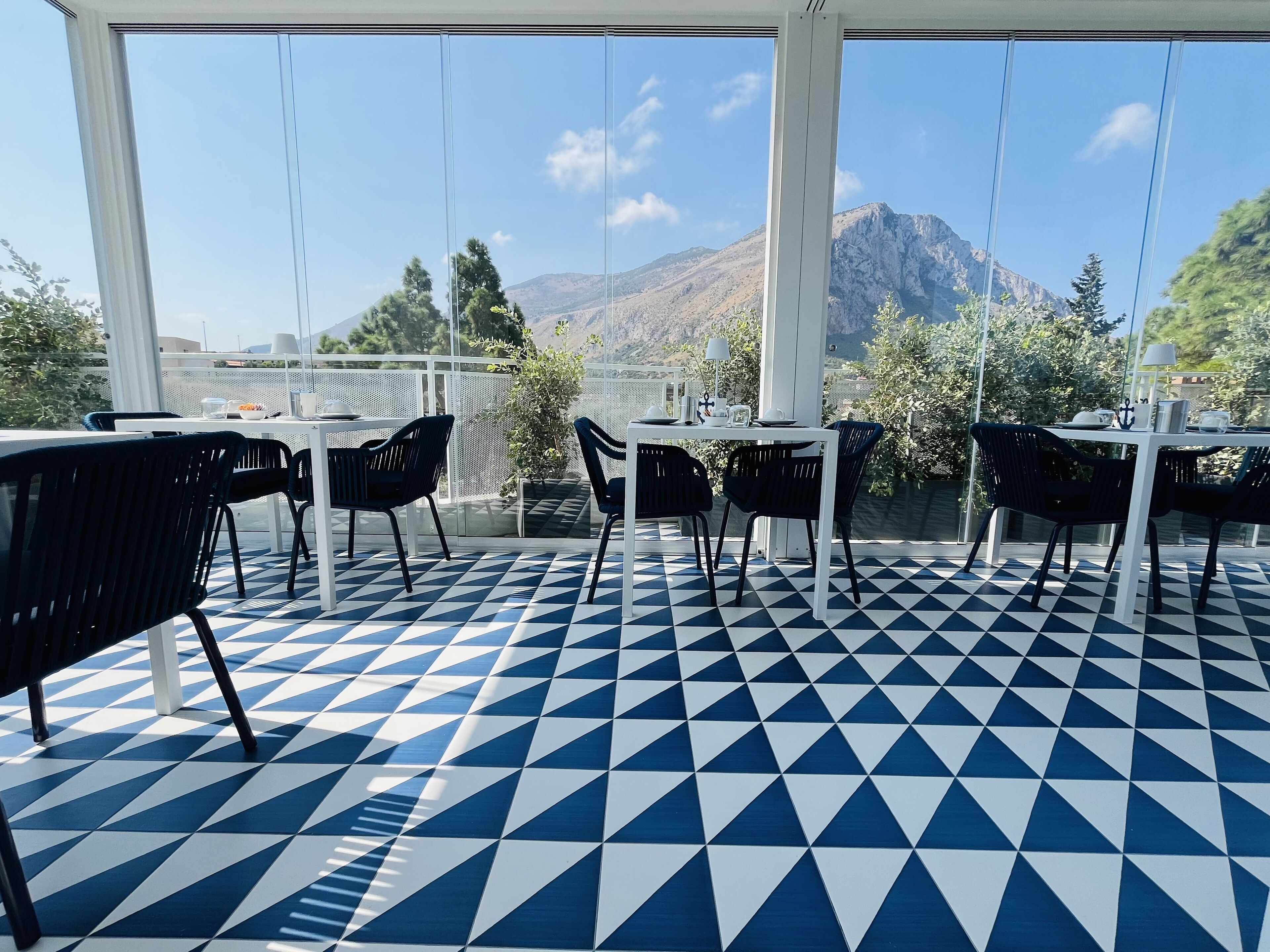 Una terraza con suelo a cuadros azul y blanco, mesas blancas y sillas negras, con vistas a un paisaje montañoso.