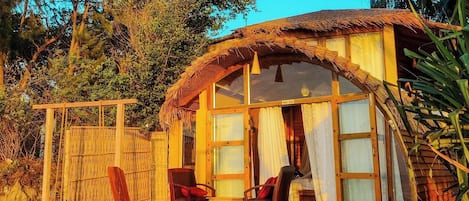 A thatched-roof hut with a wooden deck and two chairs, surrounded by trees and sand.