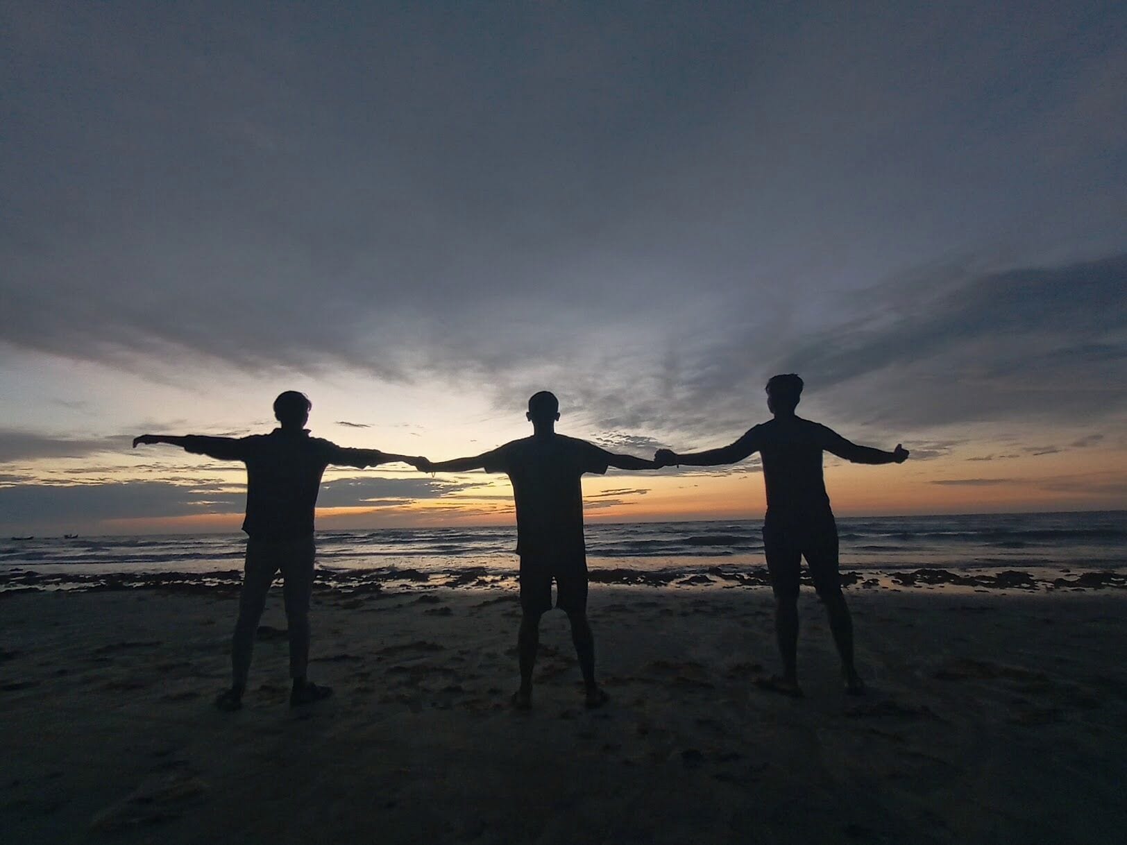 Three silhouetted figures standing on a beach with their arms outstretched, facing the ocean at sunset.