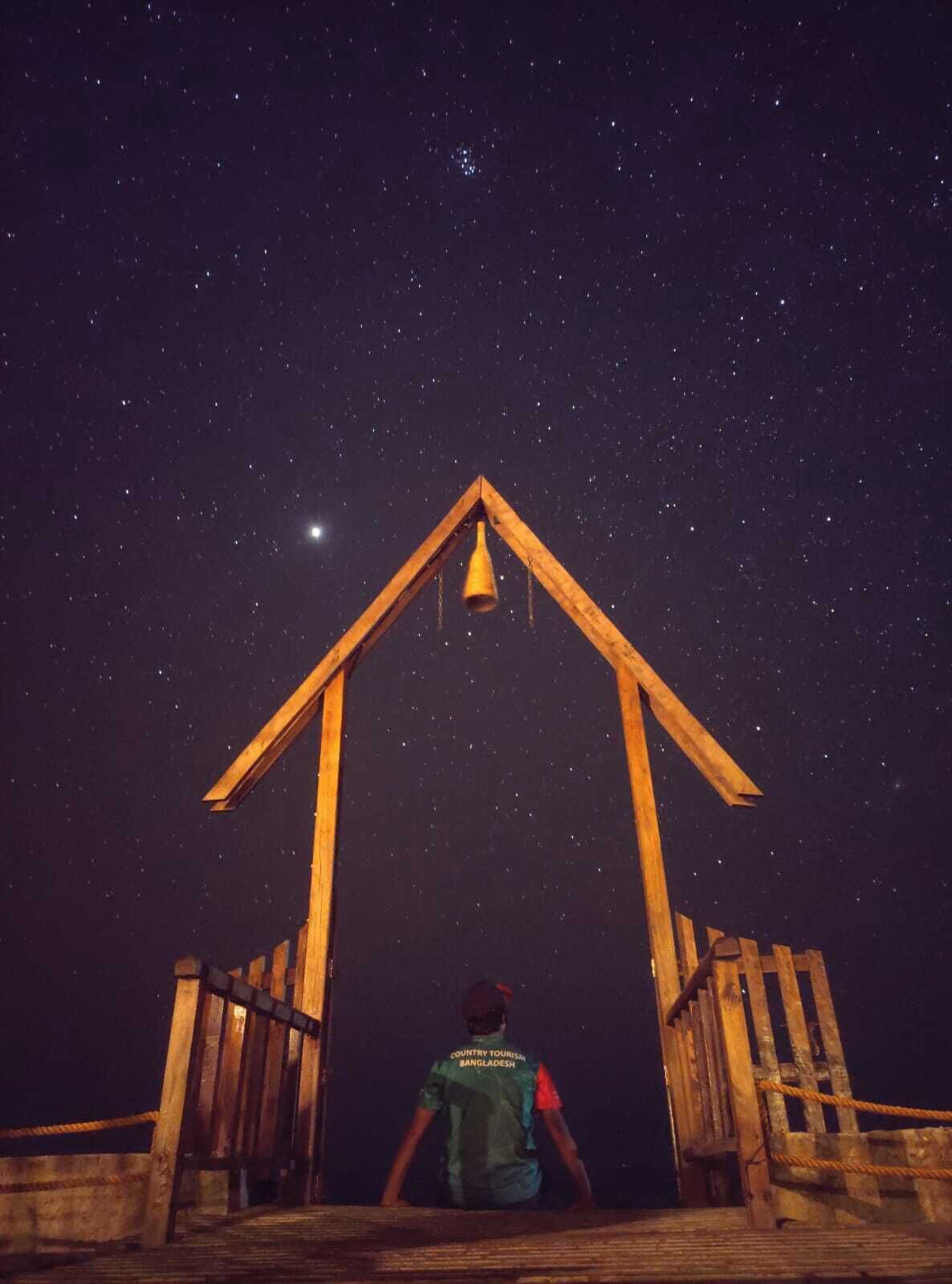 A person standing at the entrance of a wooden structure with a triangular roof, looking up at the night sky filled with stars.