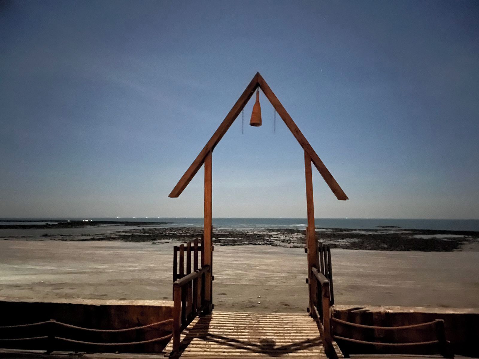 Eine Holzhütte mit einem Dreiecksdach am Strand, die zu einem Sandbereich führt.