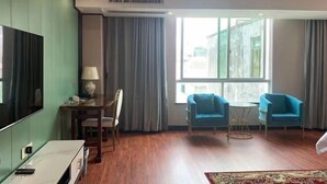 A hotel room with a large TV, a desk with a lamp, two blue armchairs, a small round table, and a large window with curtains.