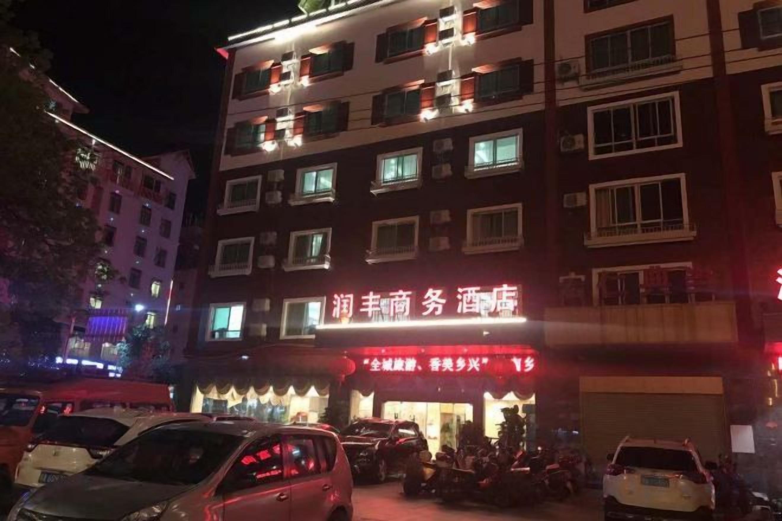 A multi-story building with illuminated signage, a parking lot with several cars, and a visible storefront with a red awning.