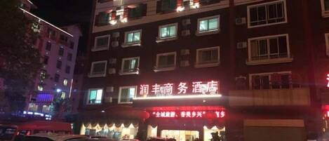 Un edificio de varios pisos con señalización iluminada, un estacionamiento con varios autos y un escaparate con toldo rojo.