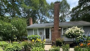Ein Wohnhaus mit Backsteinschornstein, einer Veranda und einem gepflegten Garten mit verschiedenen Pflanzen und Blumen.