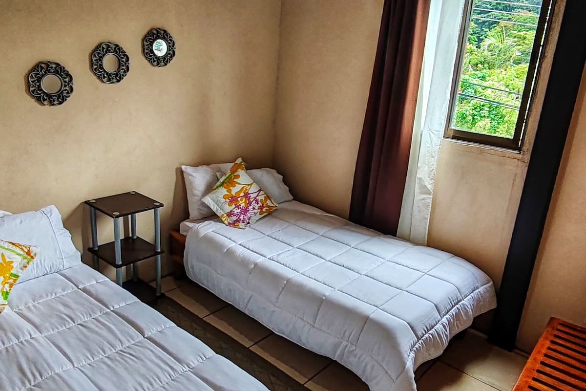 Una habitación con dos camas, una mesita y un ventanal con vistas al verdor.