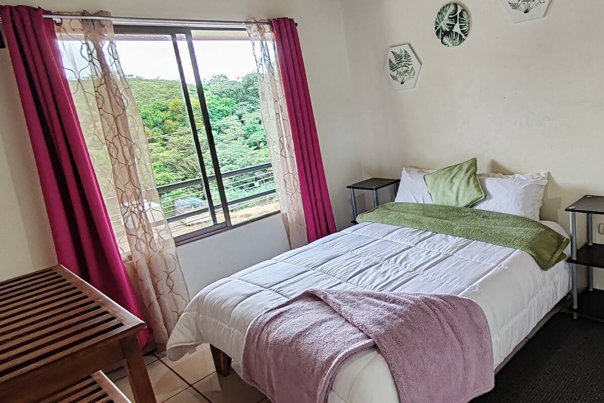 Un dormitorio con una cama, mesitas de noche, una ventana con cortinas, una puerta de madera y una pequeña mesa.