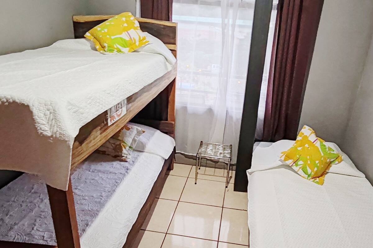 Pequeña habitación con dos camas individuales, cada una con un colchón blanco y una almohada amarilla. Hay un taburete metálico pequeño entre las camas y una ventana con una cortina ligera que la cubre parcialmente.