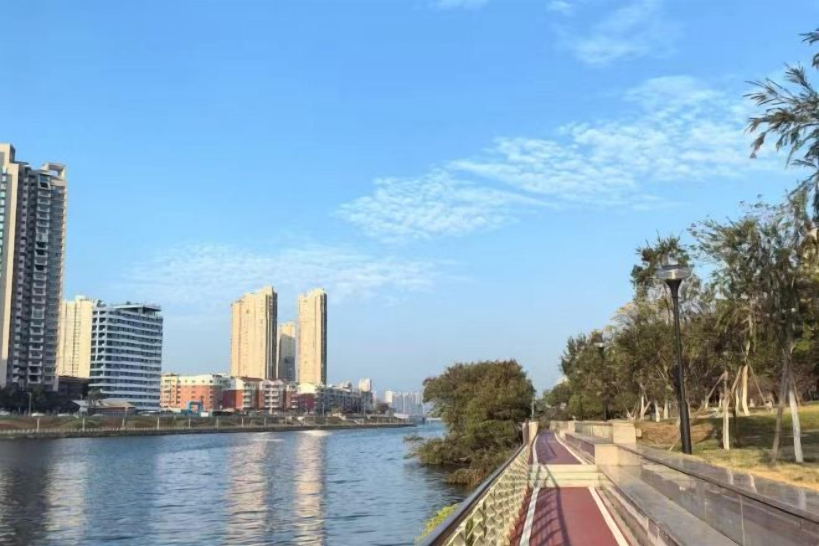 A riverside walkway with a red path, tall buildings, and trees in the background.