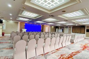 A conference room with rows of chairs, a stage with a blue backdrop, and a carpeted floor.