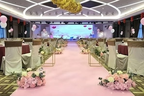 Un salón de banquetes con alfombra rosa, mesas con manteles blancos y dorados, y centros de mesa florales.