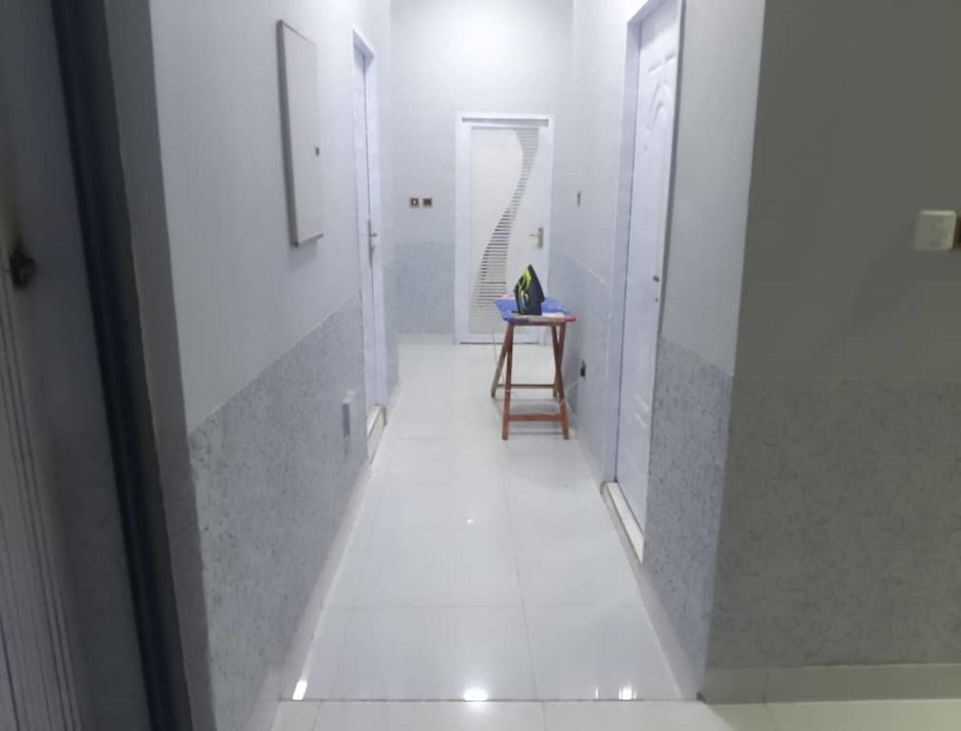 A narrow hallway with white tiled floors and walls, leading to a door at the end.