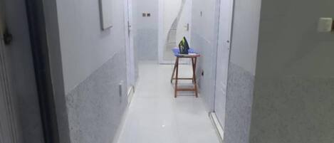 A narrow hallway with white tiled floors and walls, leading to a door at the end.
