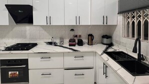 A modern kitchen with white cabinets, a black stove, a built-in oven, a coffee maker, and a window with a decorative grille. - 3 Bed Flat - Sleeps 6 - Parking - Wifi (Adentan)