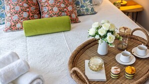 A neatly made bed with a white quilt, rolled white towels, a green pillow, a tray with tea set, and a vase with white flowers.