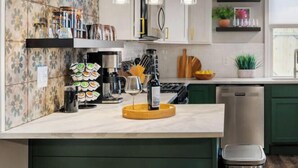 A modern kitchen with a central island, bar stools, hanging pendant lights, and a stainless steel dishwasher.
