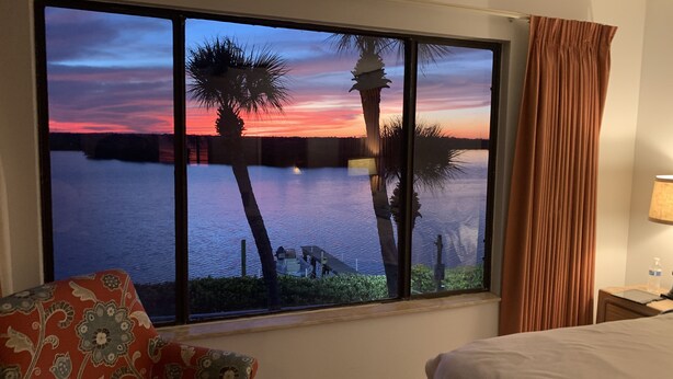 Habitación de hotel con un ventanal que ofrece vistas a un atardecer sobre el agua, una cama, una mesita de noche y una silla con cojín estampado con flores.