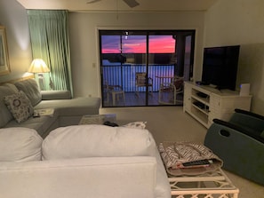 Un salon avec un canapé, un téléviseur, un balcon offrant une vue sur le coucher du soleil et une table avec des chaises.
