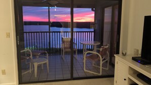 Un balcón con mesa y sillas, un televisor sobre un mueble, y vista a un atardecer sobre el agua.