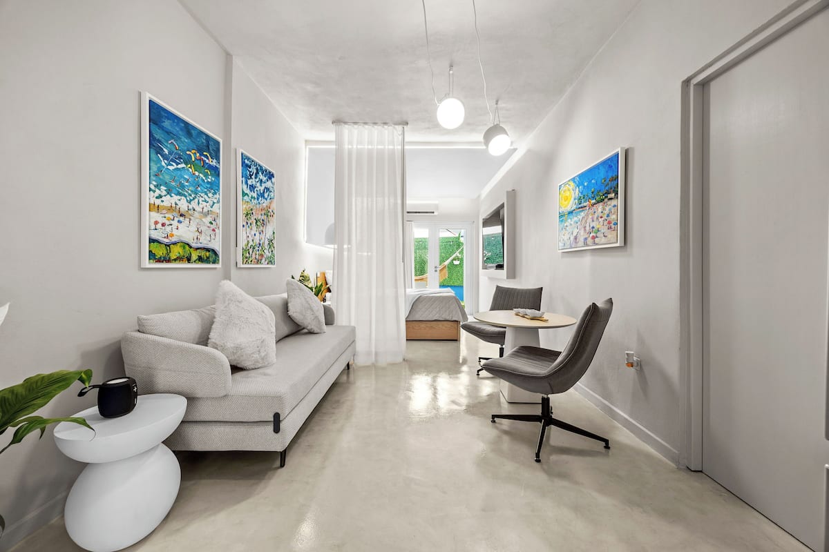 A modern living room with a sofa, a dining table, and a chair. There are paintings on the walls and a plant on the side table.