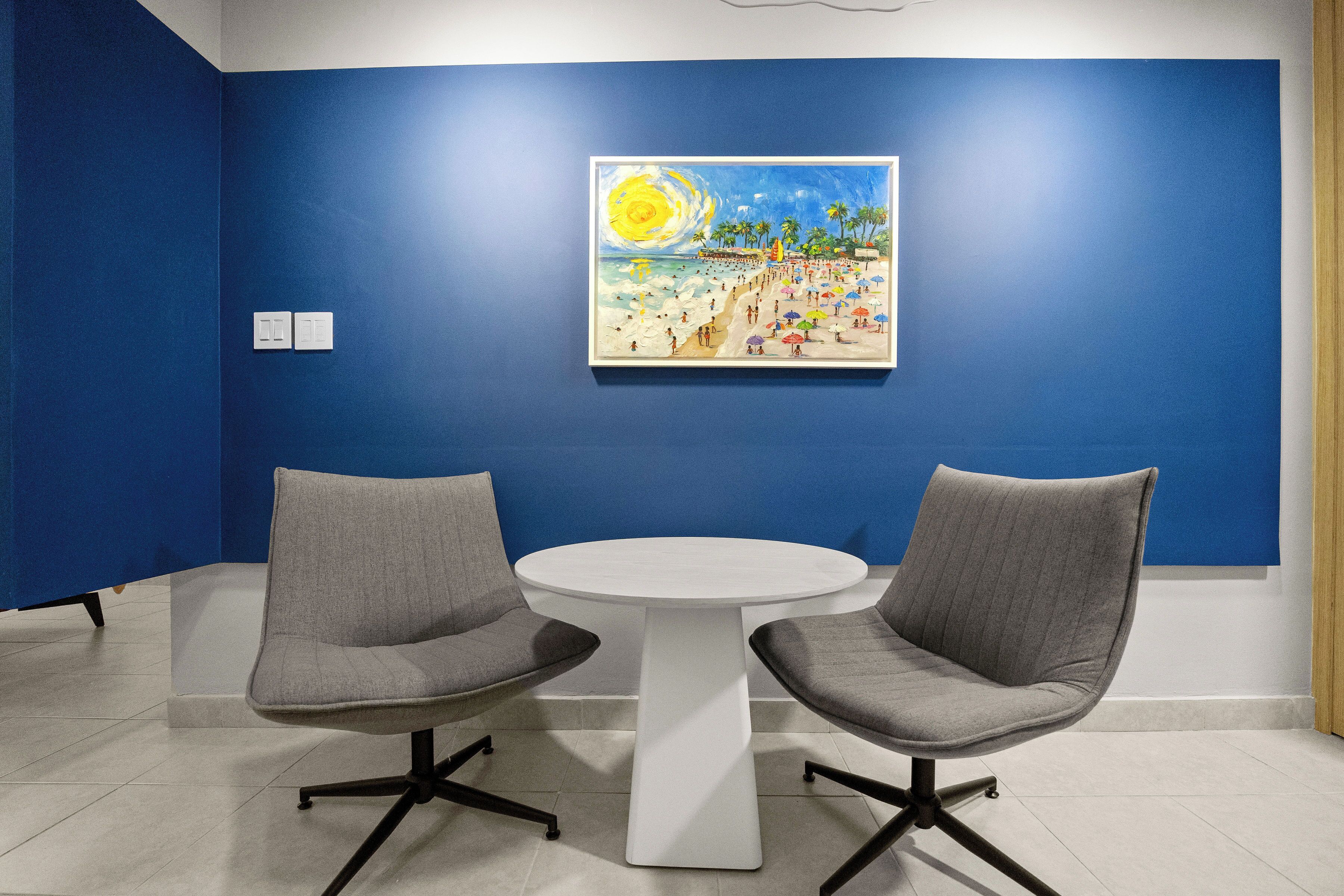 A room with two grey chairs around a white table, a painting of a beach scene on the wall, and a blue wall.