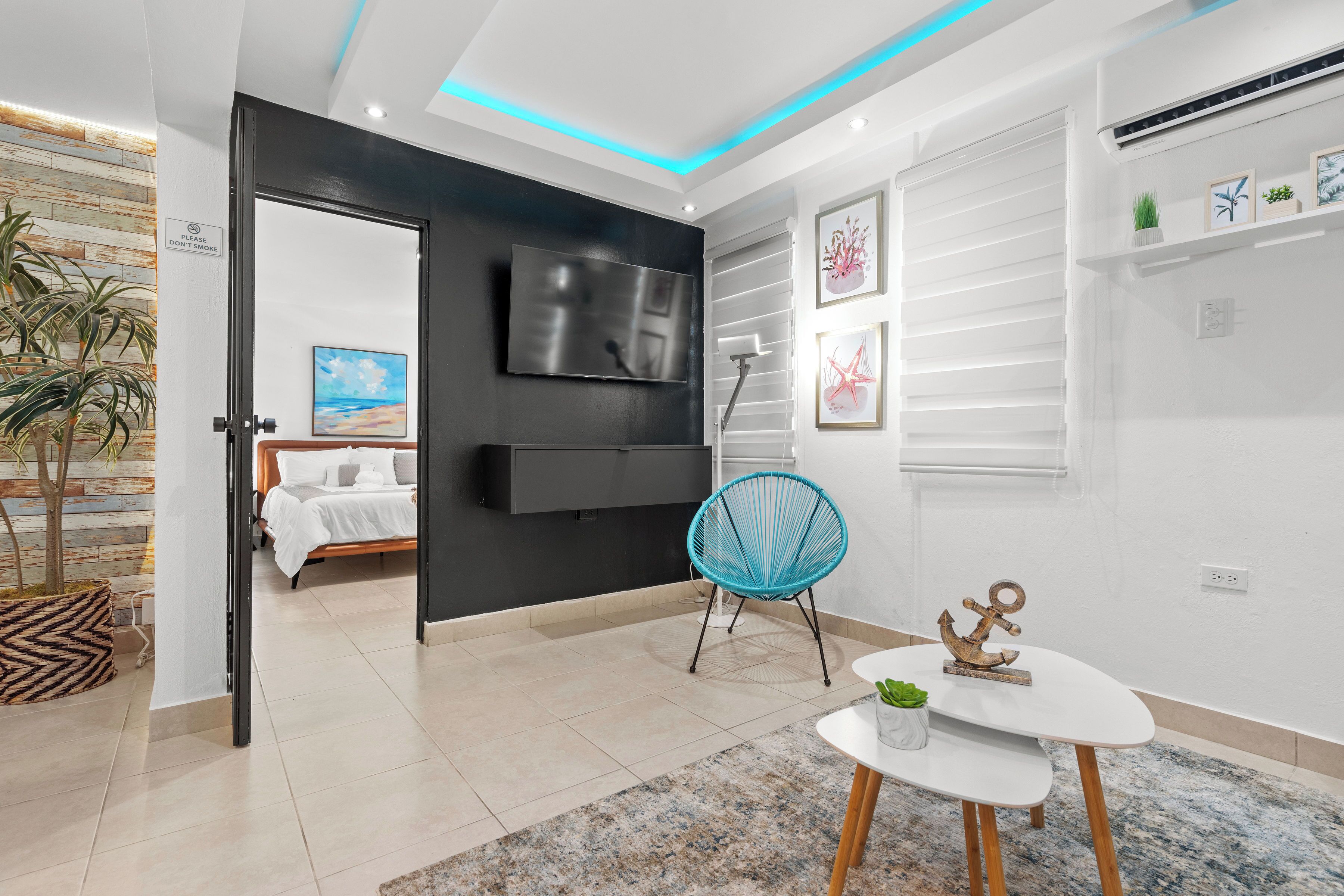 Una sala moderna con un televisor de pantalla plana, un sillón azul, una mesita con una planta y una cama al fondo.