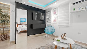 Uma sala de estar moderna com uma televisão de tela plana, uma cadeira azul, uma mesinha com uma planta e uma cama ao fundo.